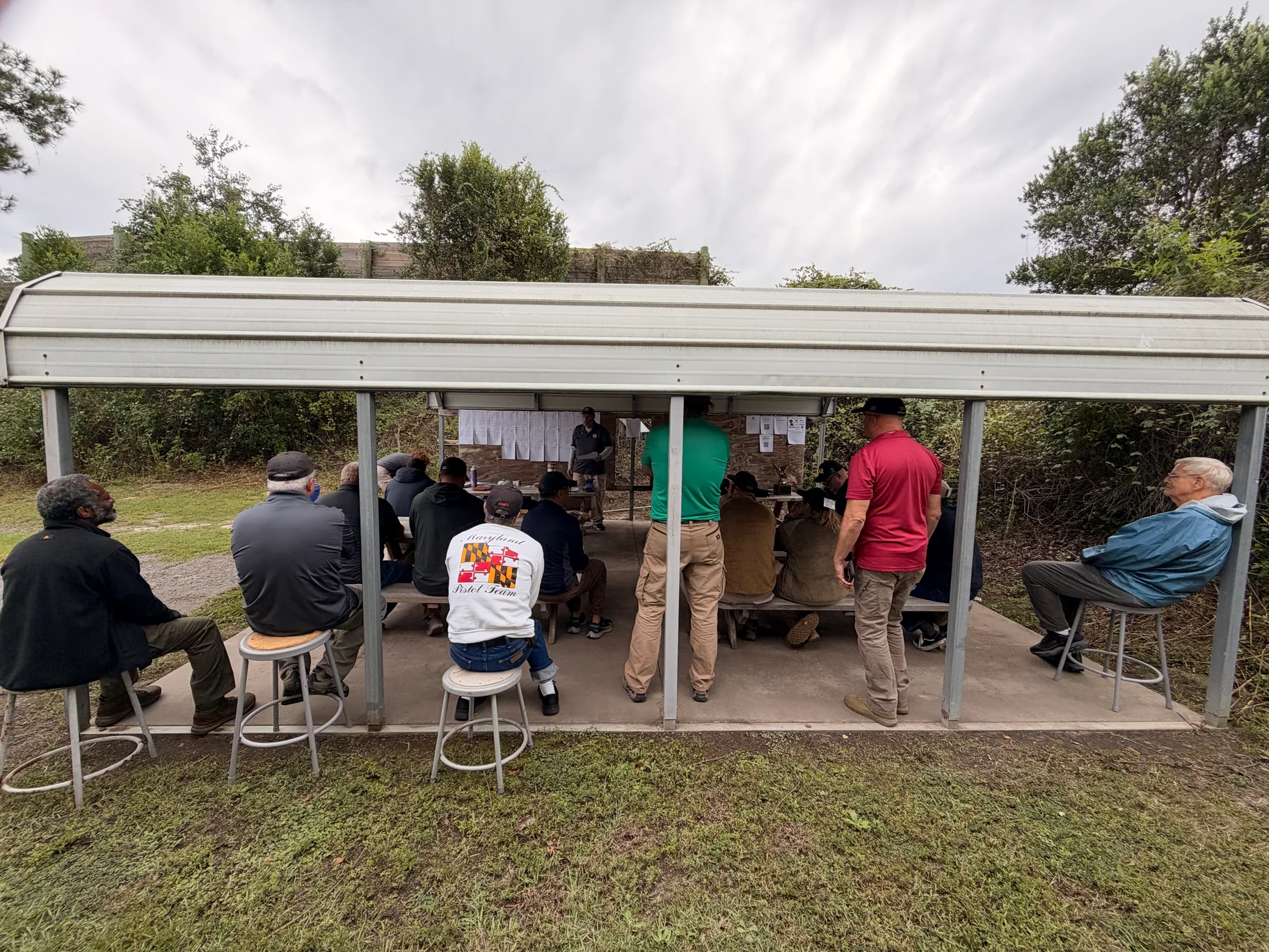Competitors gathered under a shelter beside the posted score sheets at Gateway Rifle & Pistol Club.